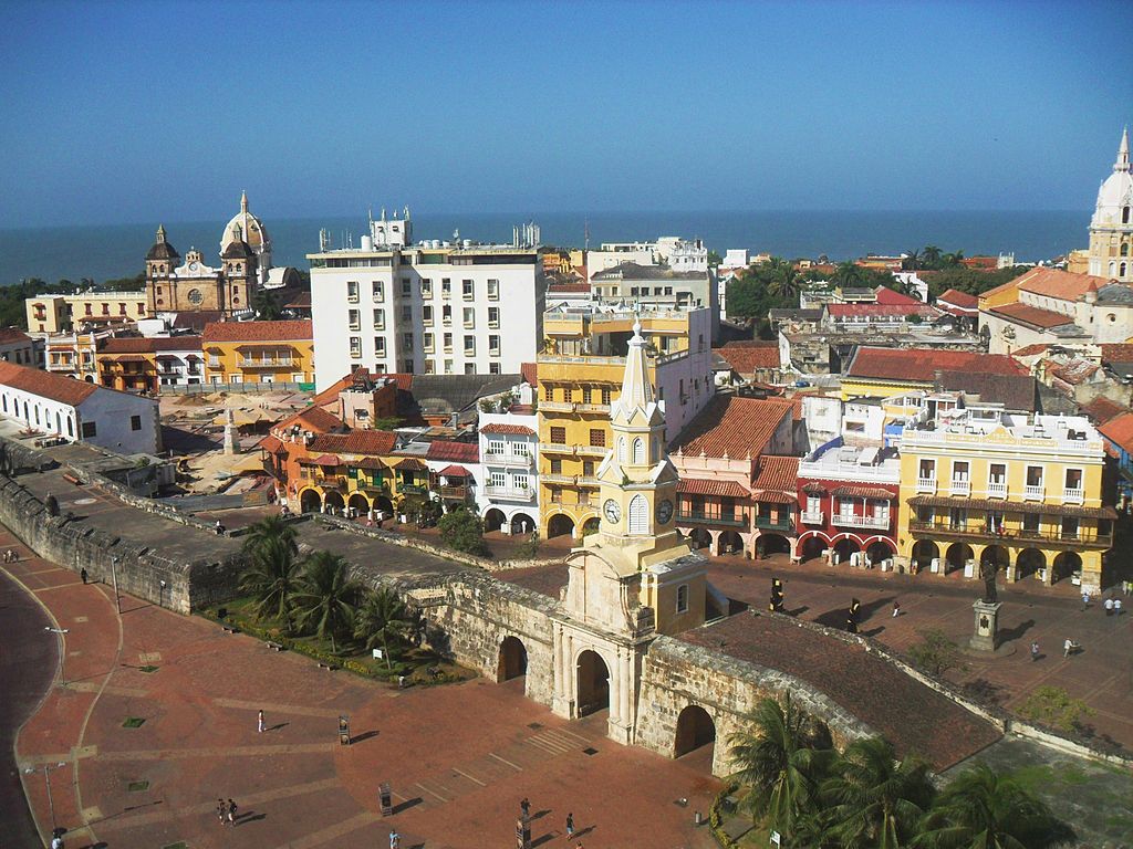 Vista desde arriba del sector antiguo de Cartagena De Indias, con la muralla defensiva y la puerta principal, que tiene una torre de reloj encima; se ven detrás unas casas tradicionales de la zona, pintadas en colores brillantes.