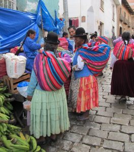 Varias mujeres vestidas al estilo indígena charlando en un mercado: llevan faldas con volantes, mantas para cargar bultos, y los sombreros negros de fieltro.
