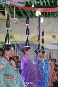 foto de cuatro mujeres haciendo la Danza de las Botellas; llevan vestidos folclóricos con faldas voluminosas que pueden menear con las manos, y encima de sus cabezas una mujer tiene dos botellas, dos mujeres tienen tres botellas, y una mujer lleva cuatro botellas apiladas verticalmente