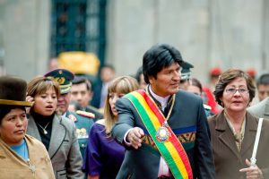 foto de Evo Morales frente a un grupo de partidarios en 2008. Morales lleva una faja representante de la presidencia de Bolivia sobre una chaqueta que combina el estilo de una chaqueta de traje moderna con elementos decorativos del tejido tradicional indígena de los Andes. A la izquierda de la foto está una mujer que lleva la manta y el sombrero tipo bombín tradicionales de las mujeres indígenas de los Andes.