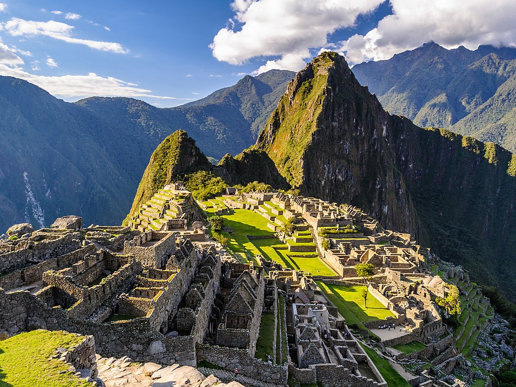 las ruinas de la Ciudadela de Machu Picchu visas desde arriba, mostrando las filas de edificios de piedra (sin techos), las terrazas verdes y los picos andinos formando el horizonte