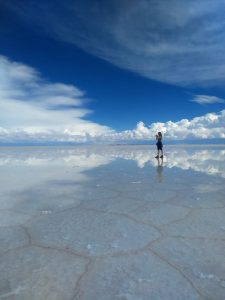 Una mujer toma fotos en el Salar de Uyuni; el cielo está reflejado en la capa fina de agua sobre la superficie del salar. Por debajo del agua también se ven las marcas hexagonales de cómo el suelo había formado placas durante la estación seca.