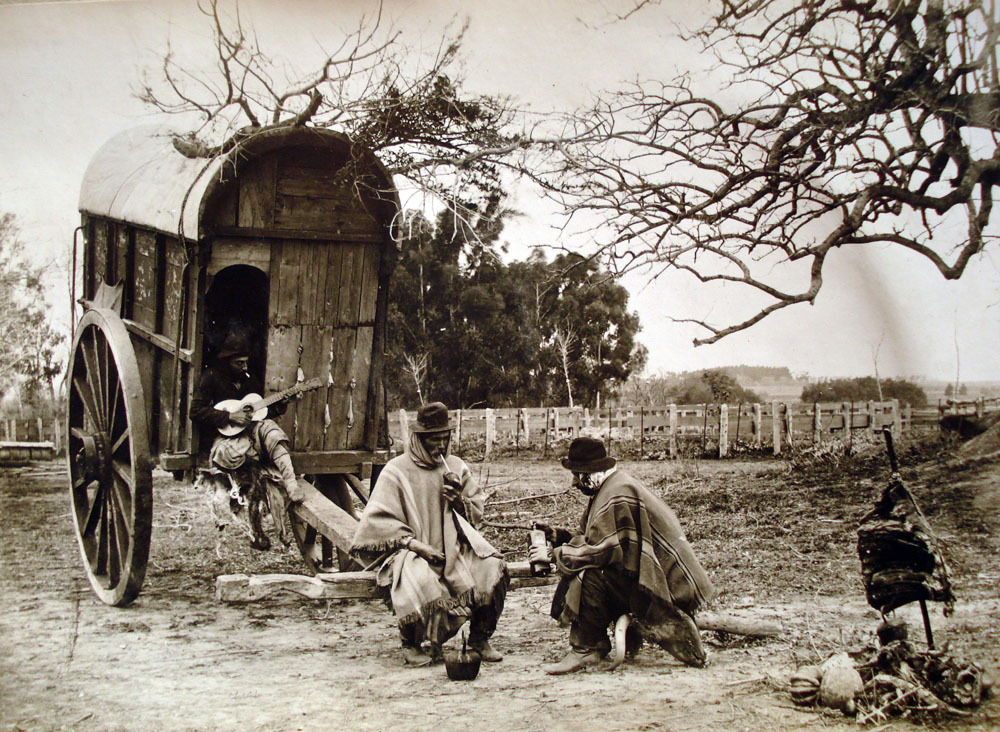 la foto muestra un gaucho sentado en una carreta de dos ruedas tocando una guitarra y dos otros gauchos que ambos llevan ponchos largos sentados en frente tomando mate. Se ven campos agrícolas detrás en la distancia.