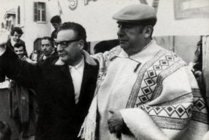 A la izquierda está el entonces presidente de Chile, Salvador Allende, llevando una chaqueta oscura y saludando al público; a la derecha está el poeta Pablo Neruda, llevando un poncho andino blanco con diseño geométrico; Neruda le da un espaldarazo a Allende.
