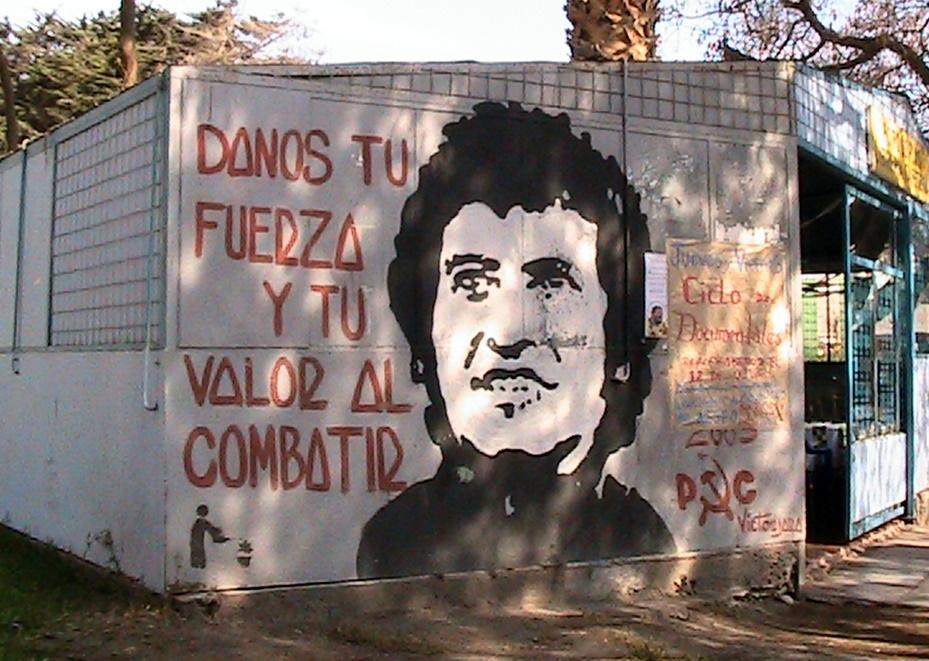 foto de un mural con el imagen de Víctor Jara y dice "Danos tu fuerza y tu valor al combatir"; los iniciales que son la firma del artista incluyen el martillo y la hoz, los símbolos del Partido Comunista